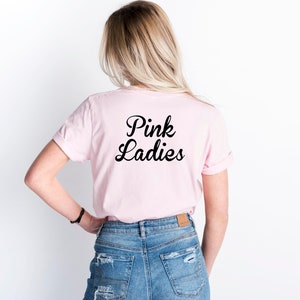 May include: A light pink t-shirt with the words "Pink Ladies" in black script. The shirt is worn by a person with blonde hair and blue jeans. The sleeves are rolled up.
