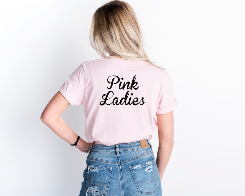 May include: A light pink t-shirt with the words "Pink Ladies" in black script. The shirt is worn by a person with blonde hair and blue jeans. The sleeves are rolled up.