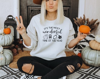 May include: Cream-colored sweatshirt with the text "It's the most wonderful time of the year" and fall-themed graphics. The image includes pumpkins, a coffee cup, a football, and a leaf. The person is holding an orange pumpkin.