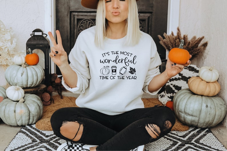 Puede incluir: Sudadera color crema con el texto "It's the most wonderful time of the year" y gr&aacute;ficos tem&aacute;ticos de oto&ntilde;o. La imagen incluye calabazas, una taza de caf&eacute;, un bal&oacute;n de f&uacute;tbol americano y una hoja. La persona sostiene una calabaza naranja.