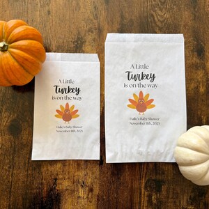 May include: Two white paper bags with the text "A Little Turkey is on the way" and a turkey graphic. An orange pumpkin and a white pumpkin are on a wooden surface. The bags are for Halle's Baby Shower, November 11th, 2025.