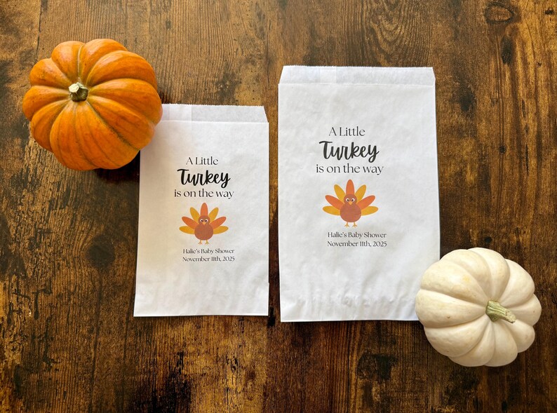May include: Two white paper bags with the text "A Little Turkey is on the way" and a turkey graphic. An orange pumpkin and a white pumpkin are on a wooden surface. The bags are for Halle's Baby Shower, November 11th, 2025.