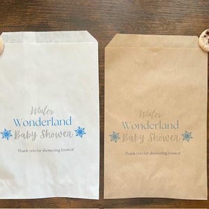 Pu&ograve; includere: Due sacchetti di carta, uno bianco e uno marrone, con la scritta "Winter Wonderland Baby Shower" in blu. Ogni sacchetto ha un disegno a fiocco di neve e un biscotto con gocce di cioccolato. Il testo "Thank you for showering Jessica!" &egrave; stampato.