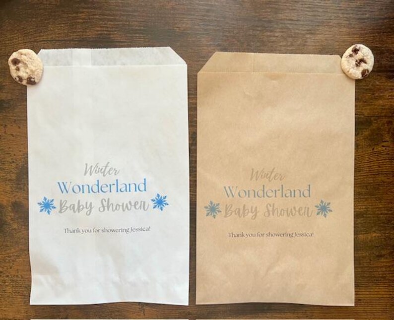 Pu&ograve; includere: Due sacchetti di carta, uno bianco e uno marrone, con la scritta "Winter Wonderland Baby Shower" in blu. Ogni sacchetto ha un disegno a fiocco di neve e un biscotto con gocce di cioccolato. Il testo "Thank you for showering Jessica!" &egrave; stampato.