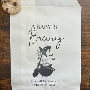 May include: A white paper bag printed with "A Baby Is Brewing" in black script, featuring a witch's hat, broom, and cauldron graphic. The bag also includes the text "Emily's Baby Shower October 7th, 2024". A chocolate chip cookie is placed on the bag.