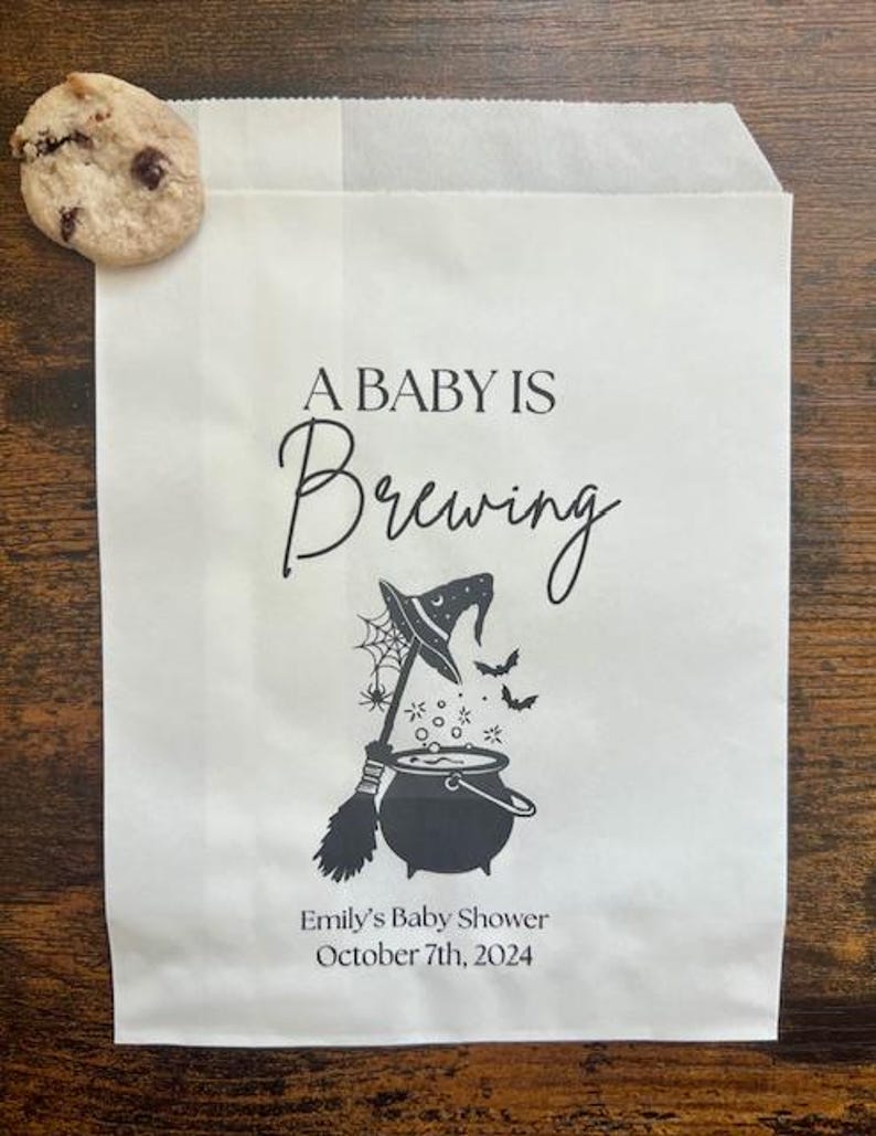 May include: A white paper bag printed with "A Baby Is Brewing" in black script, featuring a witch's hat, broom, and cauldron graphic. The bag also includes the text "Emily's Baby Shower October 7th, 2024". A chocolate chip cookie is placed on the bag.