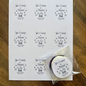 May include: A white sheet featuring eight printed rectangular labels and one circular label inside a clear plastic bag. Each label displays the black text We Created a Monster, a baby carriage icon, spider web accents, and the date October 31, 2023.
