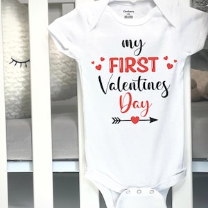 May include: White baby onesie with the text "my FIRST Valentines Day" in black and red. The design includes red hearts and an arrow with a heart. The onesie is displayed on a white crib.