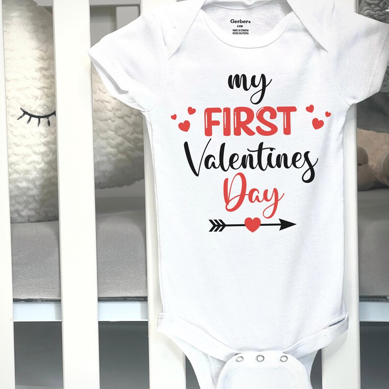 May include: White baby onesie with the text "my FIRST Valentines Day" in black and red. The design includes red hearts and an arrow with a heart. The onesie is displayed on a white crib.