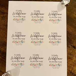 May include: A sheet of white labels with the text "A Little Wildflower is on the way" in elegant script, with floral accents. The labels are for a baby shower. Hershey's Kisses chocolates are placed near the labels.