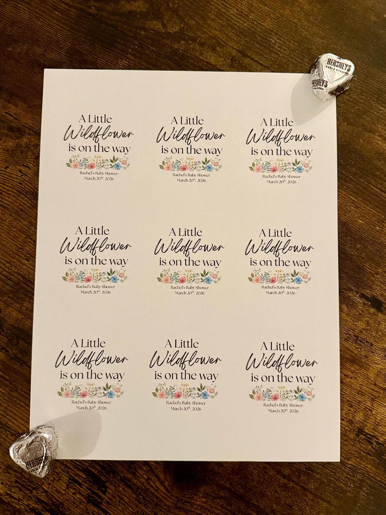 May include: A sheet of white labels with the text "A Little Wildflower is on the way" in elegant script, with floral accents. The labels are for a baby shower. Hershey's Kisses chocolates are placed near the labels.