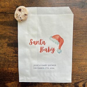 May include: A white paper gift bag with the words "Santa Baby" in red script, a watercolour Santa hat illustration, and the text "JESSICA'S BABY SHOWER DECEMBER 17TH, 2024". A chocolate chip cookie rests on the bag.