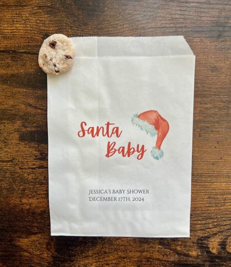 May include: A white paper gift bag with the words "Santa Baby" in red script, a watercolour Santa hat illustration, and the text "JESSICA'S BABY SHOWER DECEMBER 17TH, 2024". A chocolate chip cookie rests on the bag.