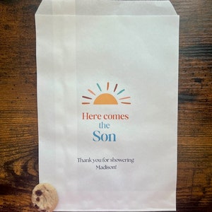 以下が含まれることがあります： カラフルな太陽のグラフィックと「Here comes the Son」の文字が入った白い紙袋。下には「Thank you for showering Madison！」と書かれています。クッキーが袋の横にあります。