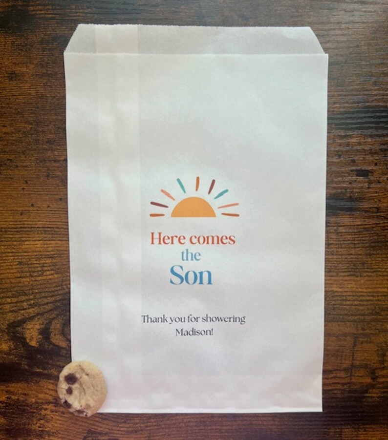 以下が含まれることがあります： カラフルな太陽のグラフィックと「Here comes the Son」の文字が入った白い紙袋。下には「Thank you for showering Madison！」と書かれています。クッキーが袋の横にあります。