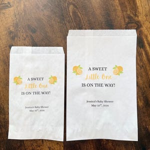 K&ouml;nnte beinhalten: Zwei wei&szlig;e Papiert&uuml;ten mit dem Text "A Sweet Little One Is On The Way!" und Orangenillustrationen. Die T&uuml;ten enthalten auch den Text "Jessica's Baby Shower May 10th, 2026."
