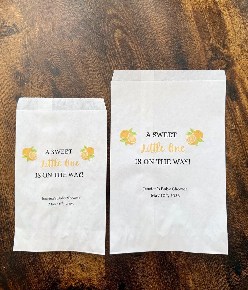 K&ouml;nnte beinhalten: Zwei wei&szlig;e Papiert&uuml;ten mit dem Text "A Sweet Little One Is On The Way!" und Orangenillustrationen. Die T&uuml;ten enthalten auch den Text "Jessica's Baby Shower May 10th, 2026."
