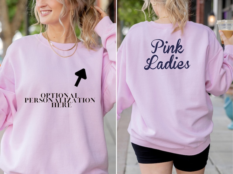 May include: A light pink sweatshirt with the text "Pink Ladies" on the back in navy blue script. The front of the sweatshirt has the text "OPTIONAL PERSONALIZATION HERE" with an arrow pointing to the text.