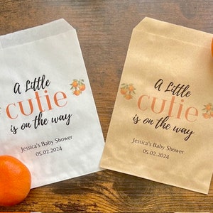 May include: Two paper treat bags, one white and one tan, with the text "A Little Cutie is on the way" and orange illustrations. The bags are for Jessica's Baby Shower on 05.02.2024. Two oranges are next to the bags.