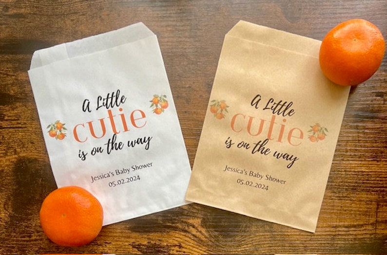 K&ouml;nnte beinhalten: Zwei Papiert&uuml;ten, eine wei&szlig;e und eine beige, mit dem Text "A Little Cutie is on the way" und Orangenillustrationen. Die T&uuml;ten sind f&uuml;r Jessicas Babyparty am 05.02.2024. Zwei Orangen liegen neben den T&uuml;ten.