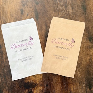 Puede incluir: Dos bolsas de papel para golosinas, una blanca y otra marr&oacute;n claro, con el texto "OUR LITTLE Butterfly IS TURNING ONE!" en escritura p&uacute;rpura. Un peque&ntilde;o gr&aacute;fico de mariposa est&aacute; encima de la palabra "Butterfly". El texto "Jessica's First Birthday April 22, 2026" est&aacute; debajo.