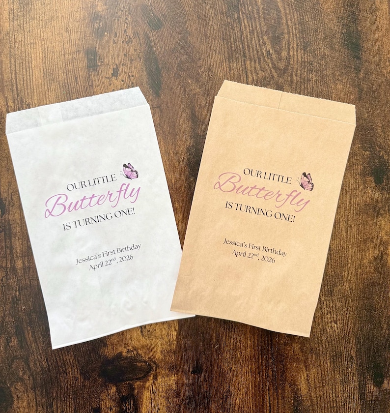 Puede incluir: Dos bolsas de papel para golosinas, una blanca y otra marr&oacute;n claro, con el texto "OUR LITTLE Butterfly IS TURNING ONE!" en escritura p&uacute;rpura. Un peque&ntilde;o gr&aacute;fico de mariposa est&aacute; encima de la palabra "Butterfly". El texto "Jessica's First Birthday April 22, 2026" est&aacute; debajo.