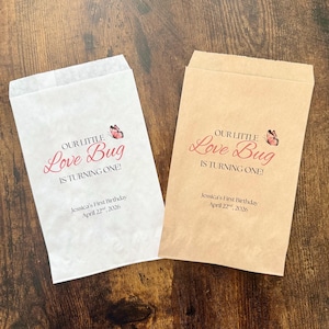 May include: Two paper treat bags, one white and one tan, with the text "OUR LITTLE Love Bug IS TURNING ONE!" in pink script. A butterfly graphic is above the text. The bottom of the bags read "Jessica's First Birthday April 22nd, 2026."