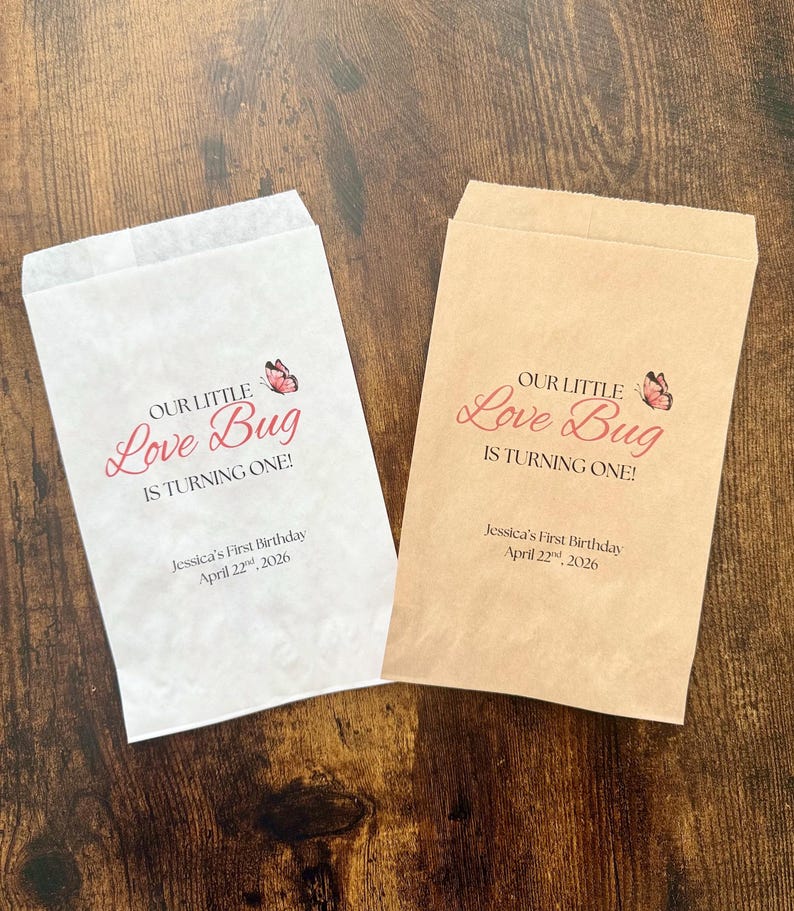 May include: Two paper treat bags, one white and one tan, with the text "OUR LITTLE Love Bug IS TURNING ONE!" in pink script. A butterfly graphic is above the text. The bottom of the bags read "Jessica's First Birthday April 22nd, 2026."