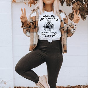May include: White t-shirt with a graphic of Michael Myers and the text "MICHAEL MYERS BUTCHER SHOP ALWAYS FRESH". The person is wearing a plaid shirt, black leggings, a tan beret, and sunglasses.