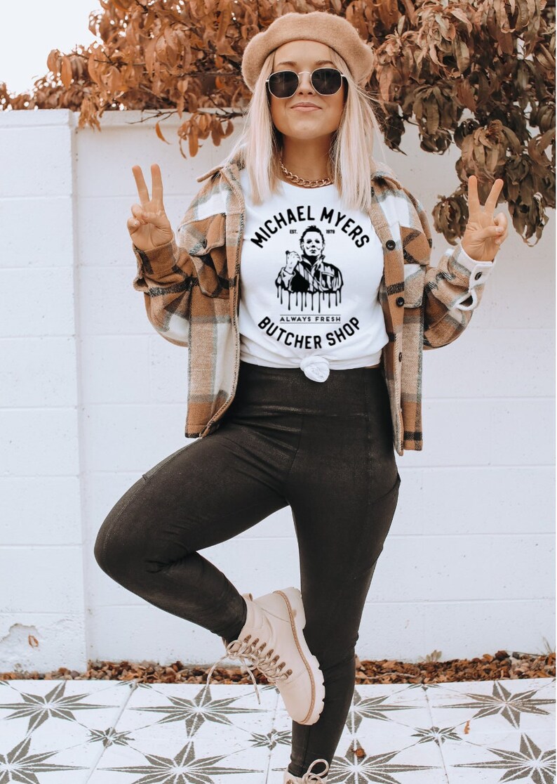 May include: White t-shirt with a graphic of Michael Myers and the text "MICHAEL MYERS BUTCHER SHOP ALWAYS FRESH". The person is wearing a plaid shirt, black leggings, a tan beret, and sunglasses.