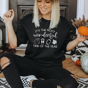 Puede incluir: Sudadera negra con texto blanco que dice "It's the most wonderful time of the year" con gr&aacute;ficos de calabaza, taza de caf&eacute;, f&uacute;tbol americano y hoja. La persona sostiene una calabaza naranja. Calabazas en el fondo.