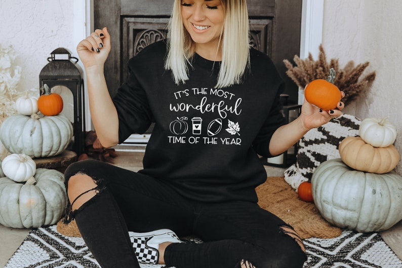 Puede incluir: Sudadera negra con texto blanco que dice "It's the most wonderful time of the year" con gr&aacute;ficos de calabaza, taza de caf&eacute;, f&uacute;tbol americano y hoja. La persona sostiene una calabaza naranja. Calabazas en el fondo.