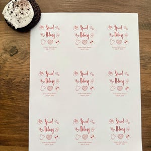 Op de afbeelding: Een wit vel met negen ronde stickers met rode tekst en afbeeldingen. Elke sticker bevat de tekst "Special Delivery" samen met iconen van een babyfles, rammelaar, rompertje, hart en wijnglazen, plus "Jessica's Baby Shower July 19th, 2026".