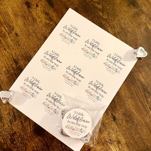 May include: A sheet of white stickers with the text "A Little Wildflower is on the way" in elegant script, surrounded by floral illustrations. A small bag contains silver charms. Hershey's Kisses are on the side.