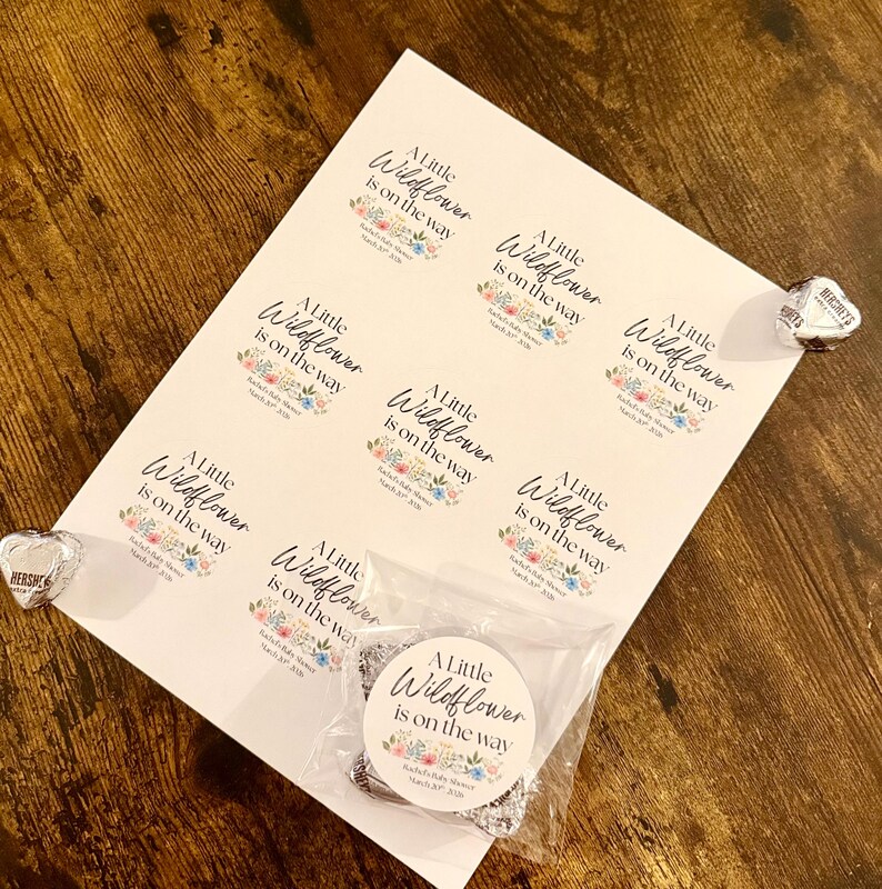 May include: A sheet of white stickers with the text "A Little Wildflower is on the way" in elegant script, surrounded by floral illustrations. A small bag contains silver charms. Hershey's Kisses are on the side.