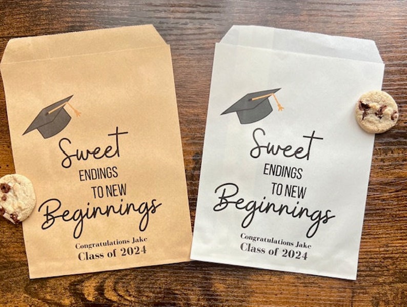 May include: Two graduation-themed treat bags, one tan and one white, with a mortarboard graphic. The bags read "Sweet Endings to New Beginnings" and "Congratulations Jake, Class of 2024". Each bag has a chocolate chip cookie.
