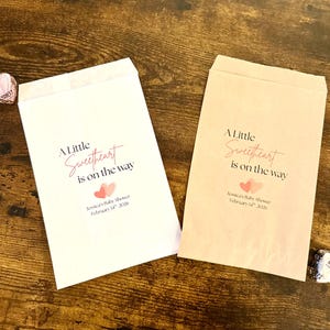 May include: Two paper treat bags with the text "A Little Sweetheart is on the way" in pink script. The bags have a baby shower theme with two pink hearts and the date February 14th, 2026.