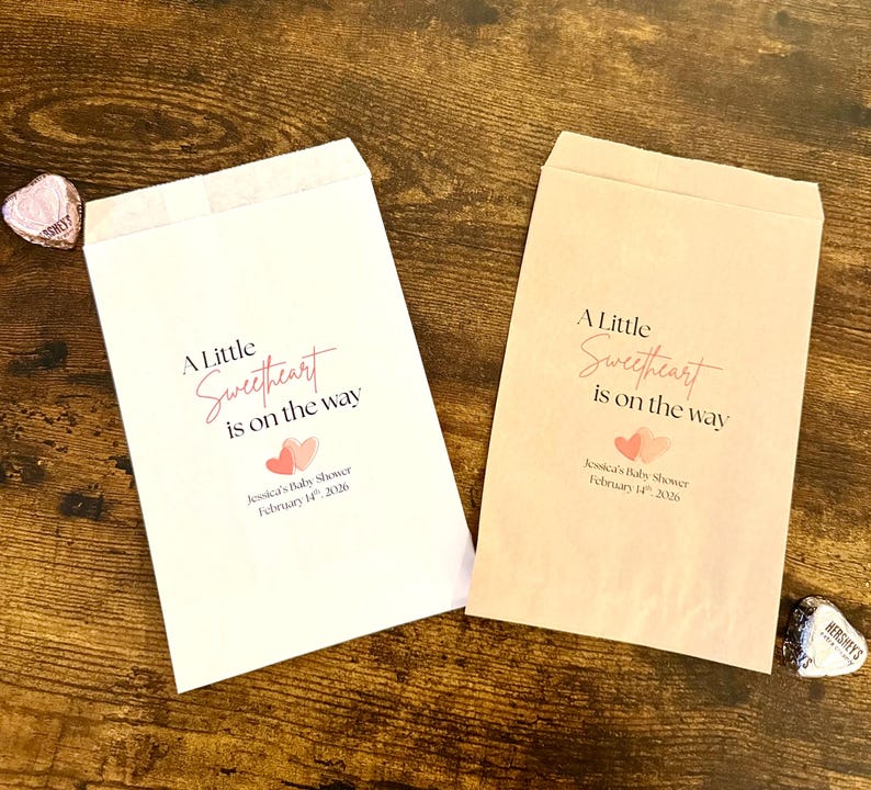 May include: Two paper treat bags with the text "A Little Sweetheart is on the way" in pink script. The bags have a baby shower theme with two pink hearts and the date February 14th, 2026.