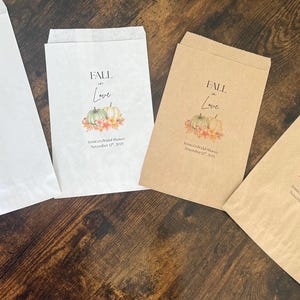 May include: Four paper favor bags with a fall theme. The bags are white and brown, with the words "FALL in Love" and a pumpkin design. Text below reads "Jessica's Bridal Shower November 17th, 2025."