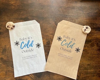 Può includere: Due sacchetti di carta, uno bianco e uno beige, con la scritta "Baby it's Cold Outside" in blu e nero. Ogni sacchetto ha un disegno a fiocco di neve e un biscotto. Il testo "Jessica's Baby Shower December 16th, 2024" è stampato sui sacchetti.