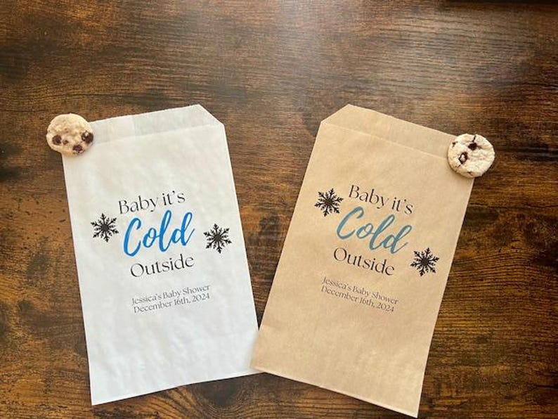 K&ouml;nnte beinhalten: Zwei Papiert&uuml;ten, eine wei&szlig;e und eine beige, mit dem Text "Baby it's Cold Outside" in Blau und Schwarz. Jede T&uuml;te hat ein Schneeflockenmuster und einen Keks. Der Text "Jessica's Baby Shower December 16th, 2024" ist auf den T&uuml;ten aufgedruckt.