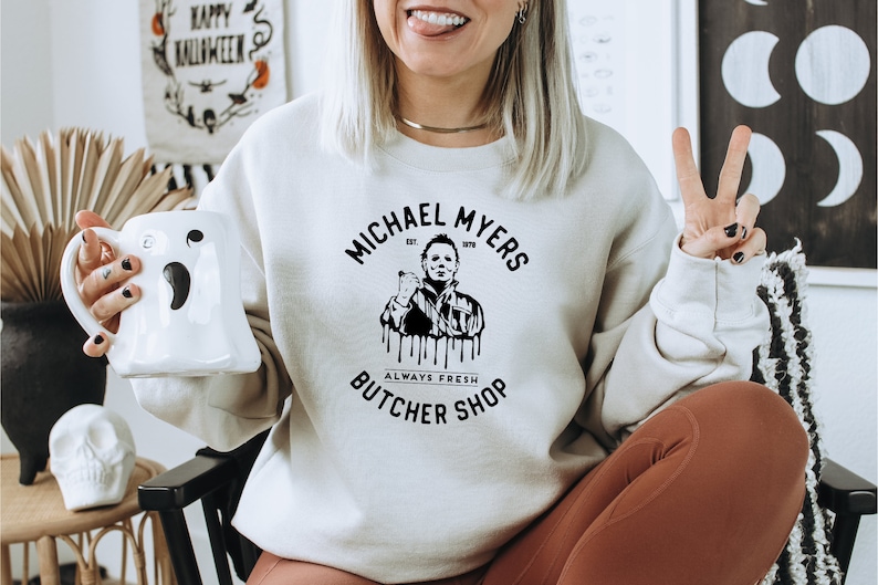 May include: Cream-colored sweatshirt with the text "MICHAEL MYERS BUTCHER SHOP" and a graphic of Michael Myers. A white ghost-shaped mug is held in the left hand. The background includes a Halloween banner and decorative items.