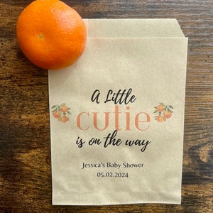 K&ouml;nnte beinhalten: Eine braune Papiert&uuml;te mit dem Text "a Little cutie is on the way" und "Jessica's Baby Shower 05.02.2024". Eine Orange liegt auf der T&uuml;te. Die T&uuml;te hat Orangenillustrationen.