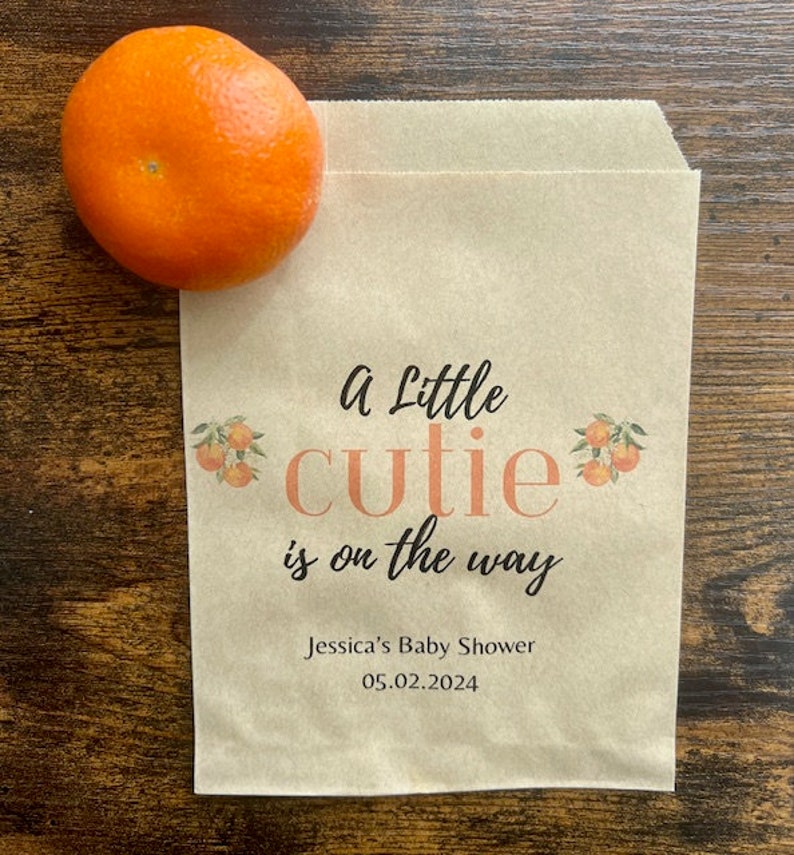 K&ouml;nnte beinhalten: Eine braune Papiert&uuml;te mit dem Text "a Little cutie is on the way" und "Jessica's Baby Shower 05.02.2024". Eine Orange liegt auf der T&uuml;te. Die T&uuml;te hat Orangenillustrationen.