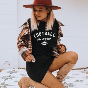 May include: Black t-shirt with the text "FOOTBALL mom" in white script and a football graphic. The shirt is worn with a brown plaid shirt and a brown hat. The model is kneeling on a patterned tile surface.