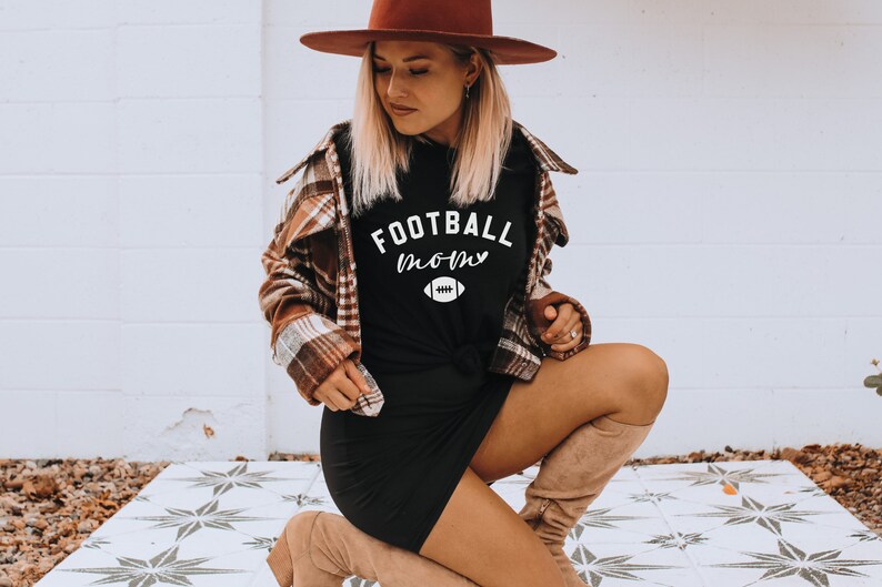 May include: Black t-shirt with the text "FOOTBALL mom" in white script and a football graphic. The shirt is worn with a brown plaid shirt and a brown hat. The model is kneeling on a patterned tile surface.