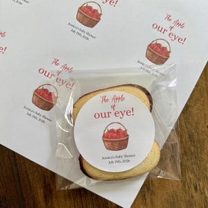 May include: A round sticker featuring a watercolor illustration of a brown woven basket filled with red apples. The text reads "The Apple of our eye!" in red script and print, followed by "Jessica's Baby Shower July 15th, 2026" in black text.