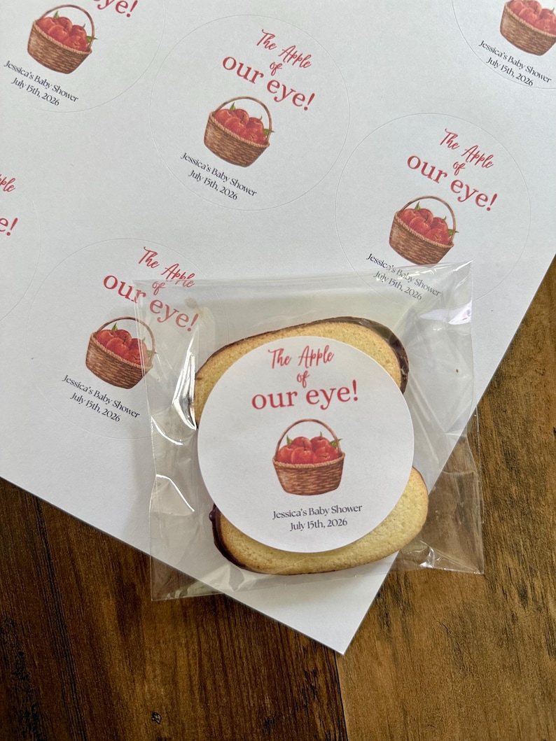 May include: A round sticker featuring a watercolor illustration of a brown woven basket filled with red apples. The text reads "The Apple of our eye!" in red script and print, followed by "Jessica's Baby Shower July 15th, 2026" in black text.