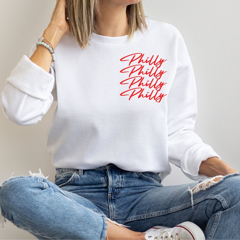 May include: White sweatshirt with the word "Philly" in red script repeated four times. The sweatshirt has long sleeves and a crew neck. The person in the image is wearing blue jeans.