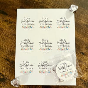 May include: A sheet of stickers and a round sticker with the text "A Little Wildflower is on the way" and floral designs. The stickers are for a baby shower. Two Hershey's Kisses are on the wooden surface.